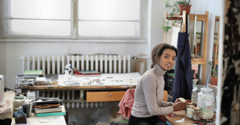 A person sits at a cluttered desk in an art studio, holding a cup amidst plants and art supplies, contemplating nonprofit challenges in the creative sector.